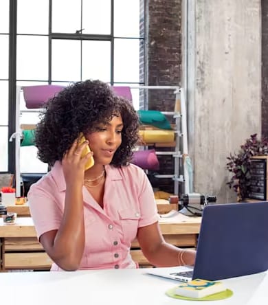 Vrouw in roze overhemd belt en werkt op laptop in creatieve werkruimte, symbool voor klantcontact en ondernemerschap