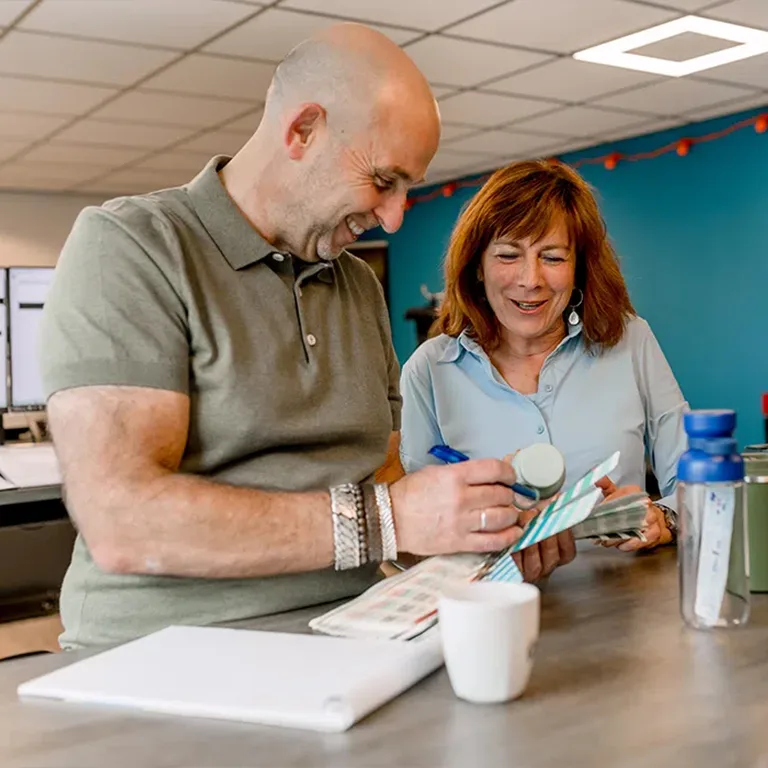 Twee collega’s overleggen lachend aan een bureau en bekijken samen kleurstalen en een drinkbeker.